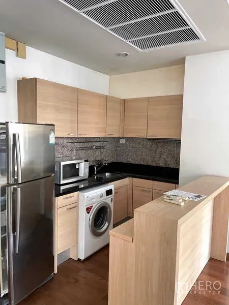 Condominium for rent in Pathum Wan, Bangkok - Fitted U-shaped kitchen with light wood cabinetry, stone countertop, fridge and washer.