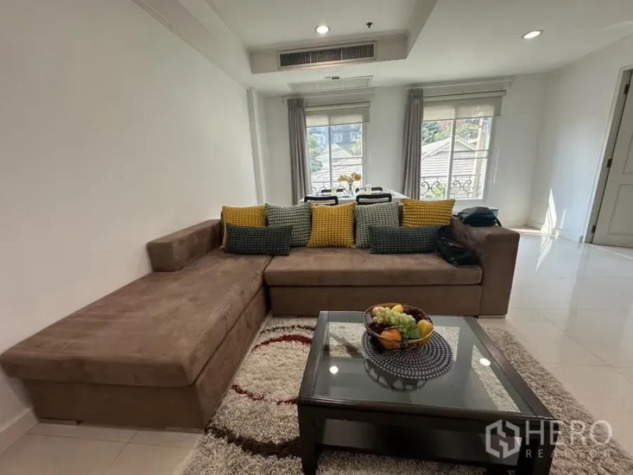 Condominium for rent - Bright living room with large L-shaped sofa, rug and glass coffee table facing a dining area by two windows.