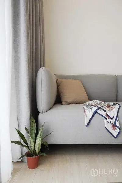Condominium for rent in Pathum Wan, Bangkok - Minimalist sofa corner with cushion, throw, and potted plant by full-height curtains.
