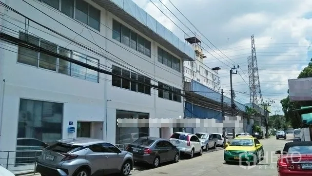 Warehouse for rent in Suan Luang, Bangkok - Street view of a white two-storey warehouse and office facade with parking and several cars.