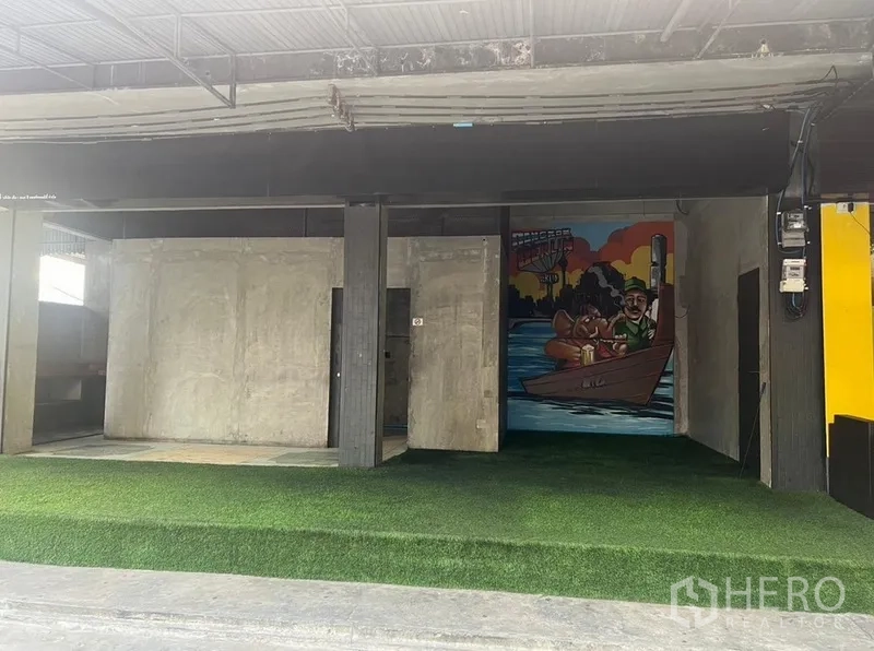 Retail Space for rent in Watthana, Bangkok - Open-front retail bay with concrete walls, artificial grass flooring, and a colorful mural beneath a metal awning.