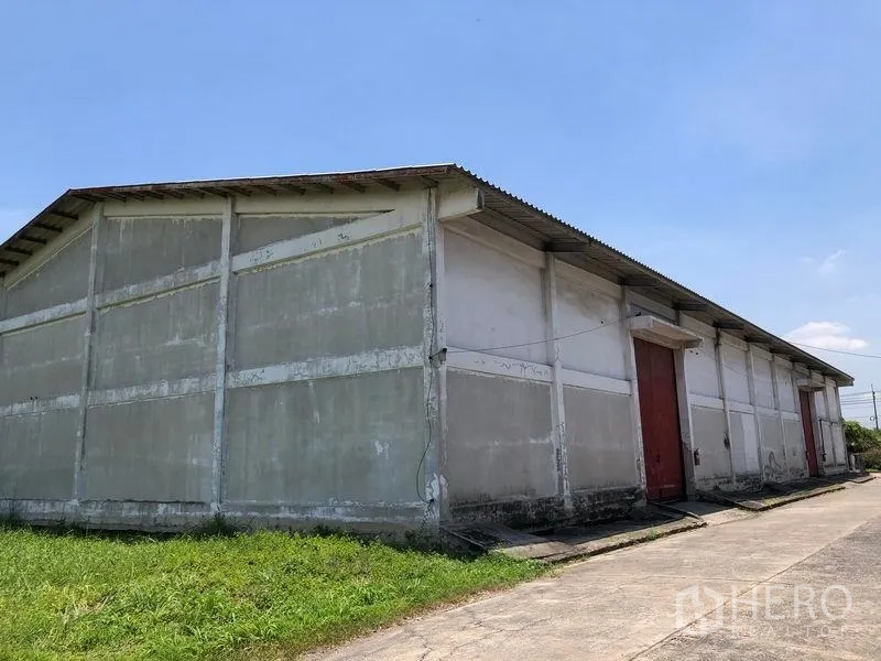 Warehouse for rent in Lat Krabang, Bangkok - Exterior view of a single-storey concrete warehouse with red sliding doors, pitched metal roof and a paved driveway under a clear sky.