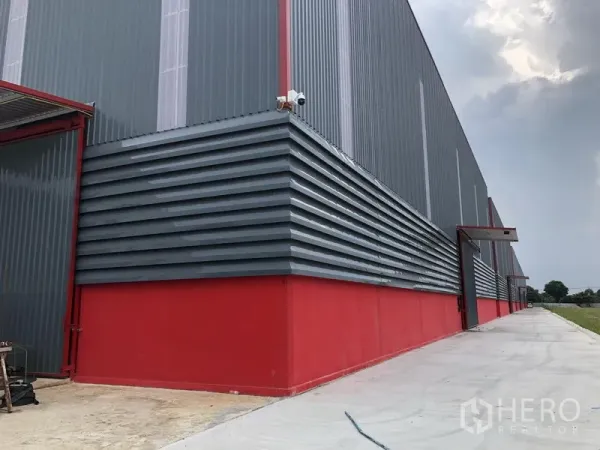 Warehouse for rent in Khlong Luang, Pathum Thani - Side corner of warehouse with corrugated metal cladding, red base, CCTV camera and covered entry.