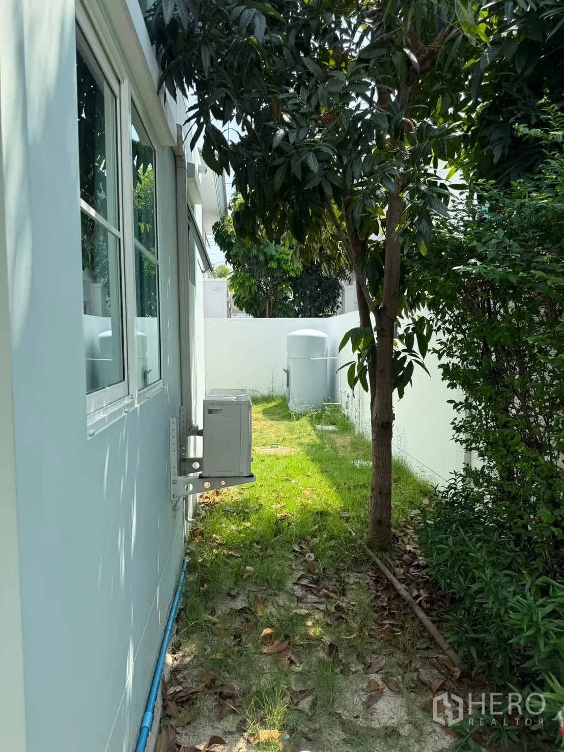 Detached House for rent in Bang Phli, Samut Prakan - Side garden with grass, mature tree, shrubs, and an exterior air-conditioning unit along the wall.
