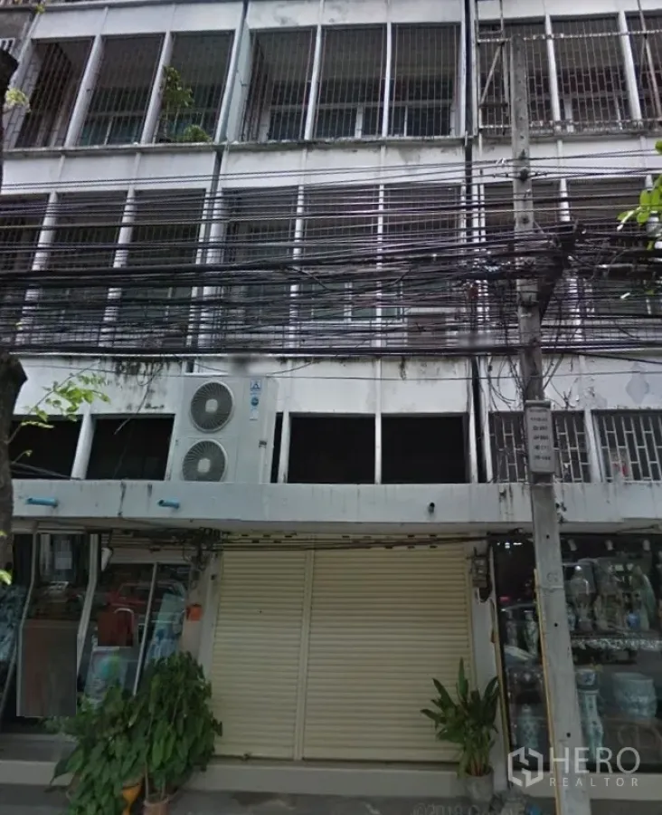 Shophouse for rent in Phra Khanong, Bangkok - Street view of a multi-level shophouse exterior with metal-grille windows, closed roller shutter, glass storefront, air-conditioning units, and dense overhead cables.