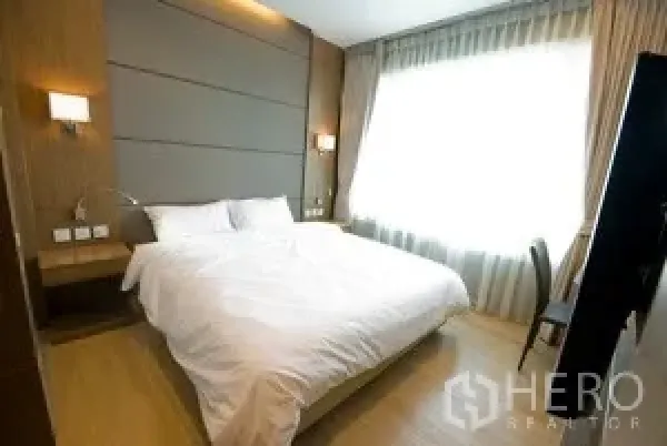 Condominium for rent in Khlong Toei, Bangkok - Master bedroom with king bed, padded headboard, bedside sconces, and large curtained window.