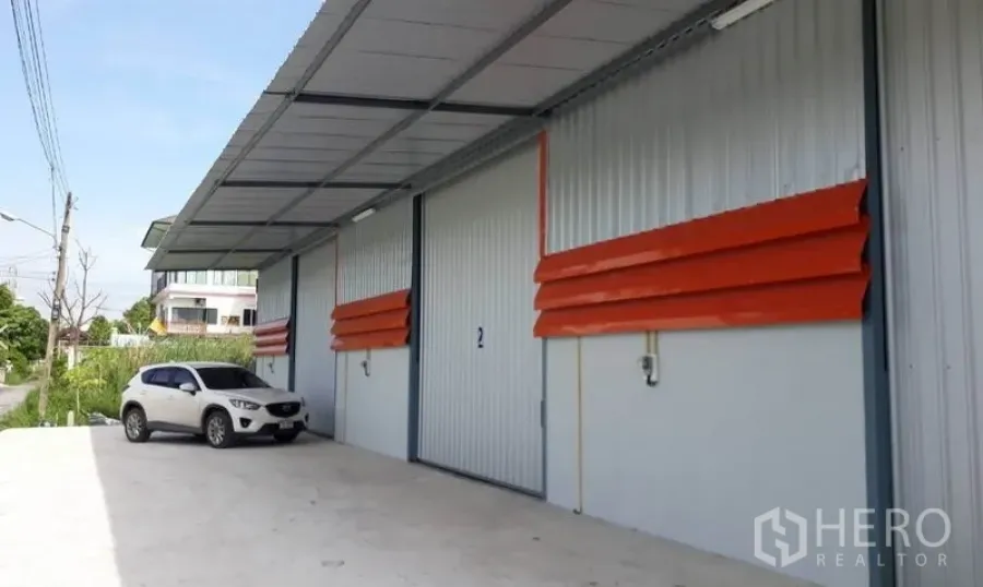 货栈 出租在Mueang Pathum Thani, Pathum Thani - 仓库外观，宽大型卷闸门与有棚装卸区，铺装车道与停车位，位于巴吞他尼市。