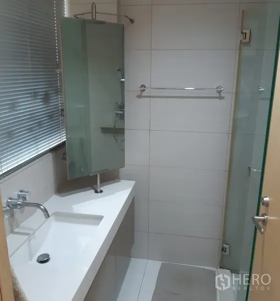 Condominium for rent in Khlong Toei, Bangkok - Contemporary bathroom with long vanity, white basin, window blinds and glass shower enclosure.