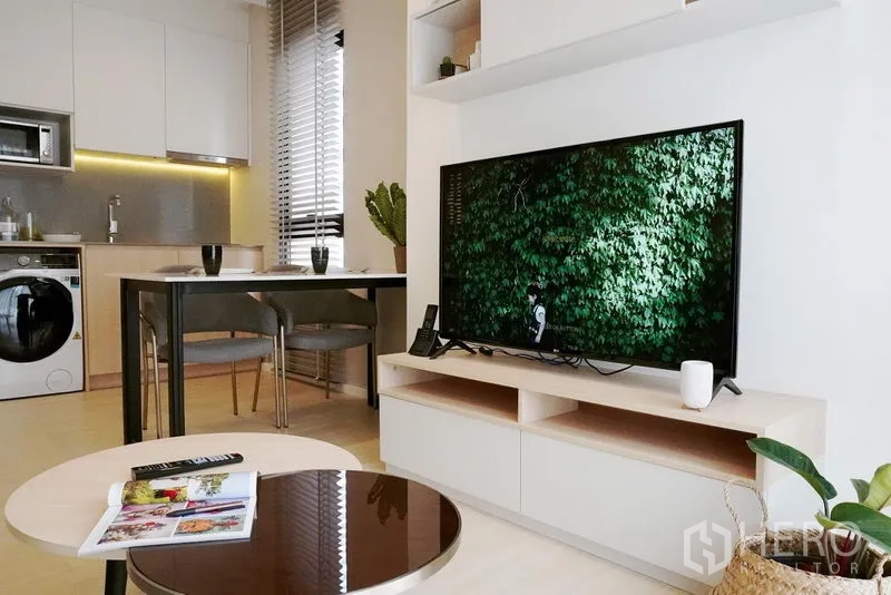 Condominium for rent in Pathum Wan, Bangkok - TV wall unit and round coffee table with sleek kitchen and dining set in the background.