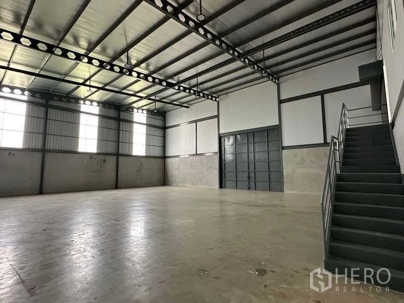 Warehouse for rent in Samut Prakan - Spacious warehouse interior with high steel roof, concrete floor, tall windows and metal staircase.