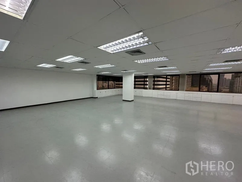 Office Space for rent in Watthana, Bangkok - Open-plan office interior with bright fluorescent lights, white ceiling, built-in low cabinets under wide windows and city views.