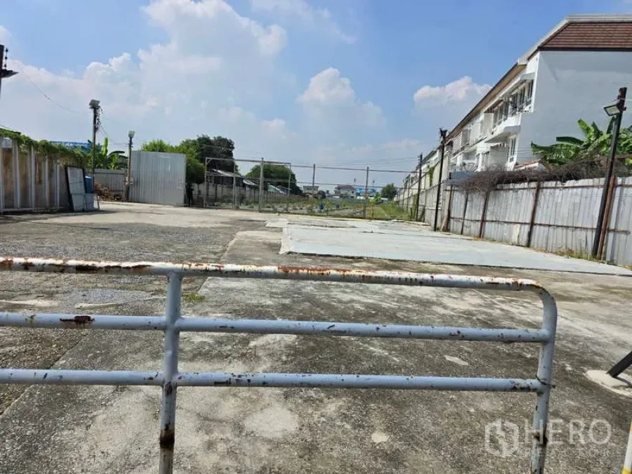 Land for rent in Prawet, Bangkok - Ground-level view of the gated, paved land plot with wide concrete yard, fenced boundaries and neighboring townhomes under a clear sky.