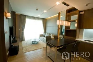 Condominium for rent in Khlong Toei, Bangkok - Dining space with pendant lights facing the living area and compact kitchen, floor-to-ceiling curtains in view.