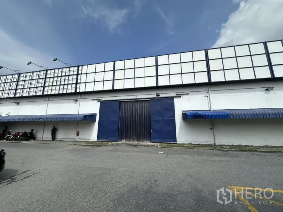 Warehouse for rent in Phra Khanong, Bangkok - Front exterior of white industrial warehouse with blue sliding door, awnings and wide parking apron under a clear sky.