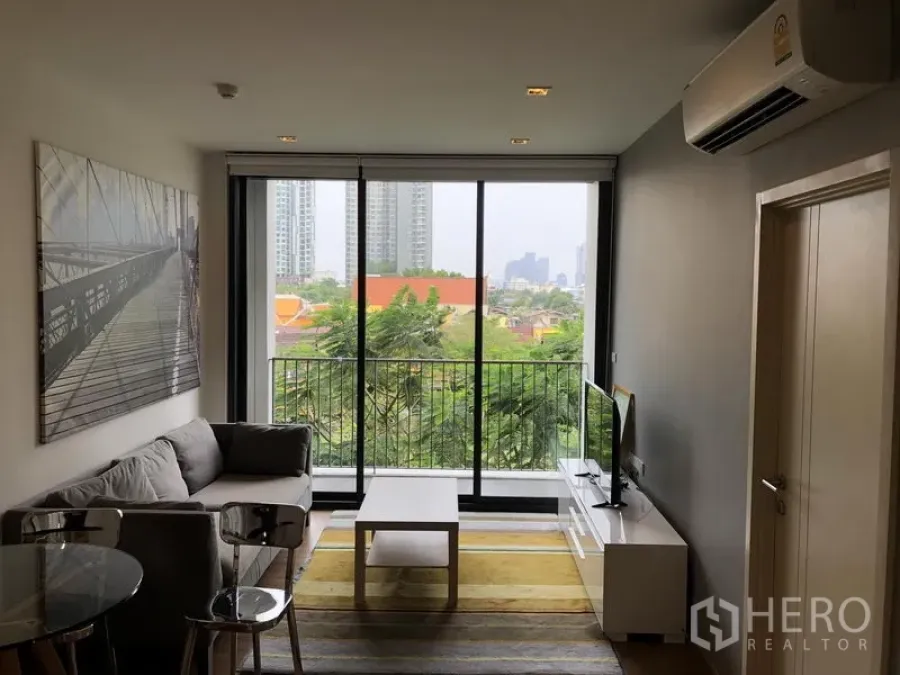 Condominium for rent in Watthana, Bangkok - Bright living room with floor-to-ceiling windows opening to a balcony with leafy city views.