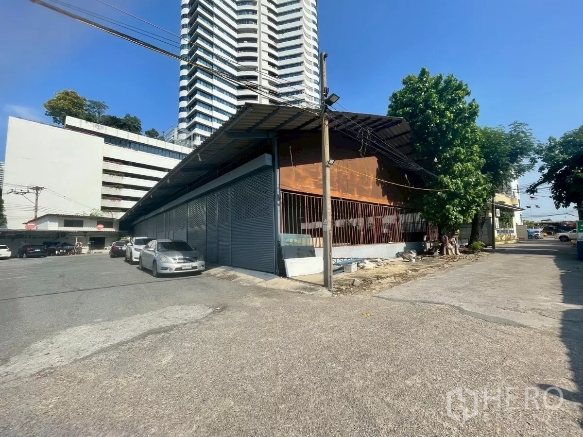Warehouse for rent or sale in Yan Nawa, Bangkok - Corner view of a single-storey warehouse with metal roof and street access beside high-rise buildings in the Rama 3 area.