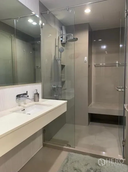 Condominium for rent in Khlong Toei, Bangkok - Modern bathroom with wide vanity, large mirror, and glass-enclosed shower with bench.
