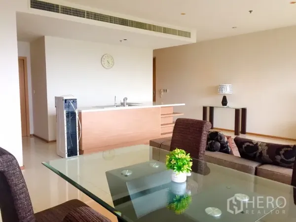 Condominium for rent in Khlong Toei, Bangkok - Open-plan living with glass-top dining table, brown chairs, sofa and a minimalist kitchen island under concealed air-conditioning.