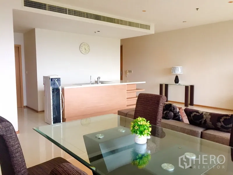 Condominium for rent in Khlong Toei, Bangkok - Open-plan living with glass-top dining table, brown chairs, sofa and a minimalist kitchen island under concealed air-conditioning.