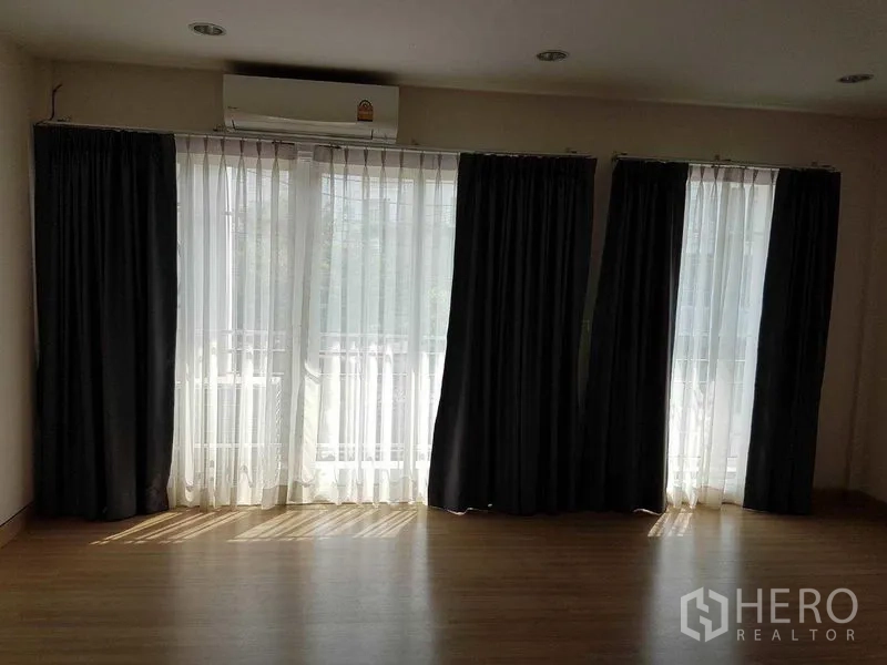 Townhouse for rent in Bang Na, Bangkok - Sunlit living area with floor-to-ceiling windows, sheer curtains and dark drapes over wood flooring in a Bang Na townhouse.