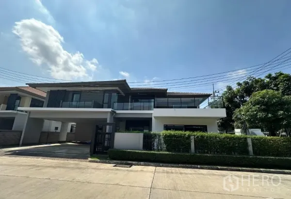 Detached House for sale in Lam Luk Ka, Pathum Thani - Daytime front view of the two-storey house with hedge fence and a covered carport.
