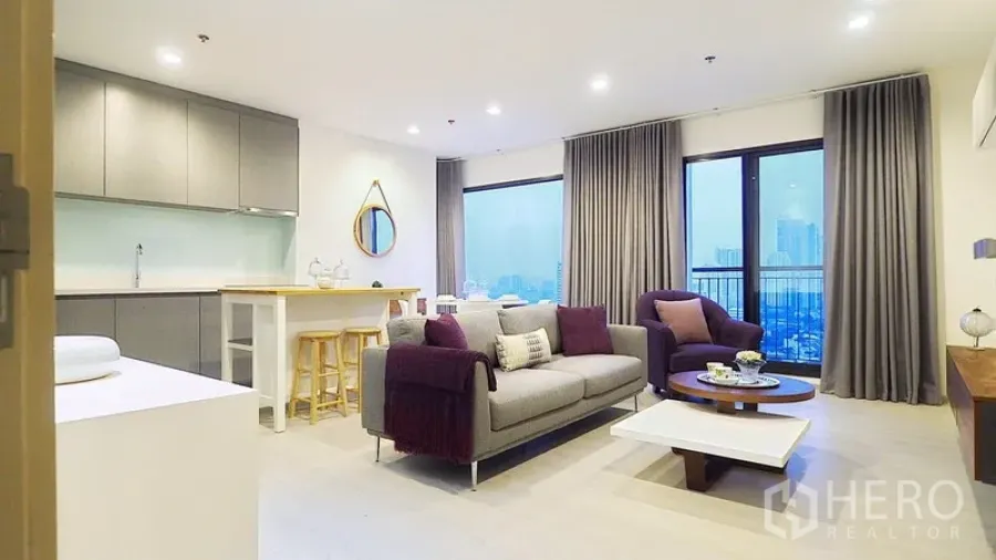 Condominium for rent or sale in Khlong Toei, Bangkok - Open-plan living room with sofa and armchair, balcony city view, and kitchen with island breakfast bar.