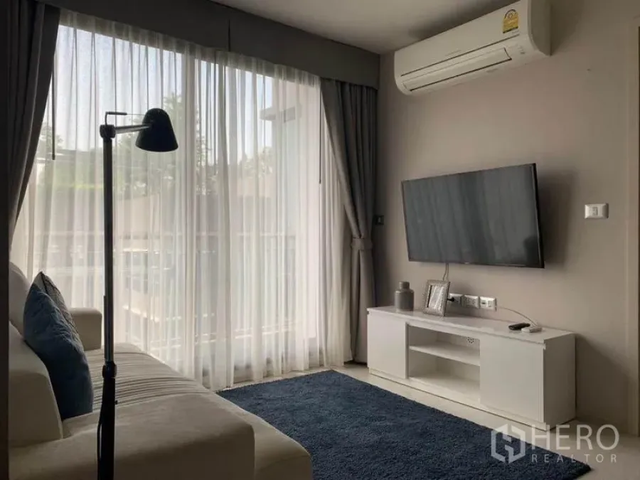 Condominium for rent in Khlong Toei, Bangkok - Living room with sofa, floor lamp, wall-mounted TV, and balcony behind sheer curtains.