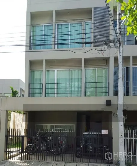 Townhouse for rent in Suan Luang, Bangkok - Modern three-storey townhouse at Noble Cube Suan Luang with sliding glass windows, slim balconies, and a gated covered parking area on a quiet residential lane.