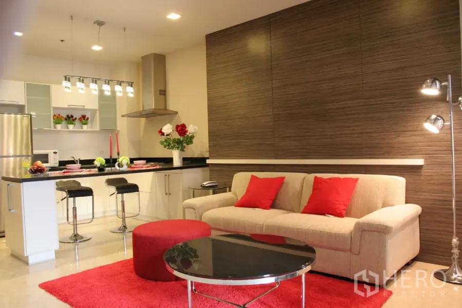 Condominium for rent in Khlong Toei, Bangkok - Open-plan living room with beige sofa, red accents, and a modern kitchen with island and pendant lights.
