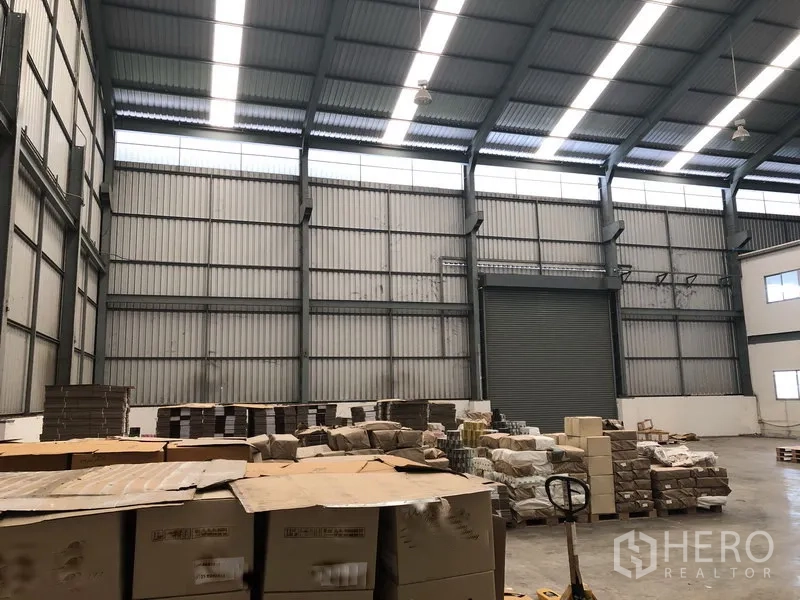 工厂 出租在Khlong Luang, Pathum Thani - 带天窗与钢梁的仓库大厅，堆放托盘，配有大型卷帘门。