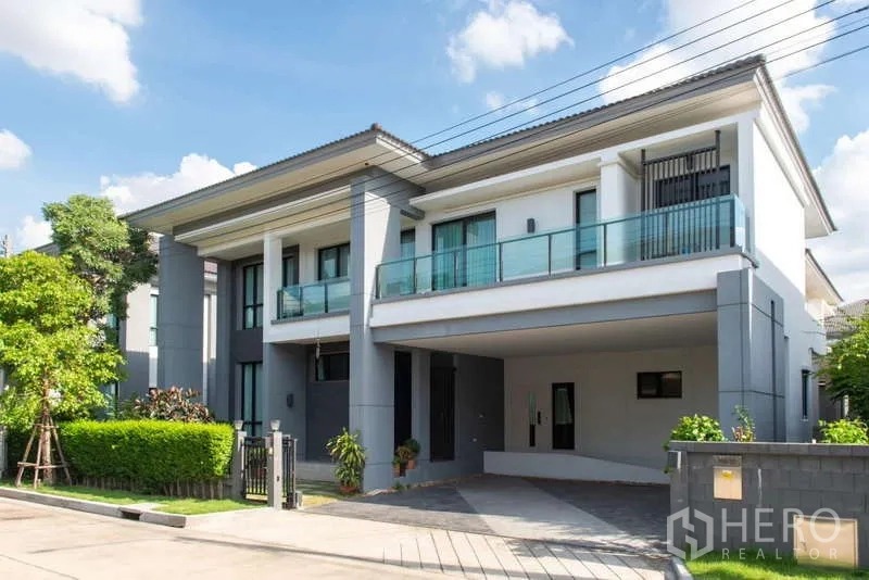 Detached House for rent or sale in Kanchanaburi - Modern two-storey detached house with glass balcony, carport, and manicured hedges under a blue sky.