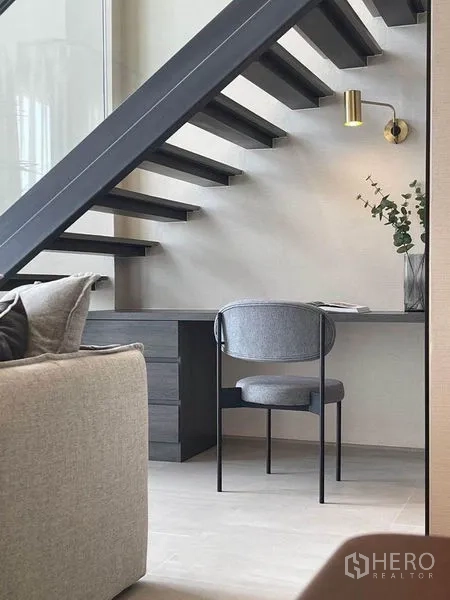 Condominium for rent in Pathum Wan, Bangkok - Under-stair home office nook with sleek desk, grey chair, brass wall sconce and open-riser staircase.