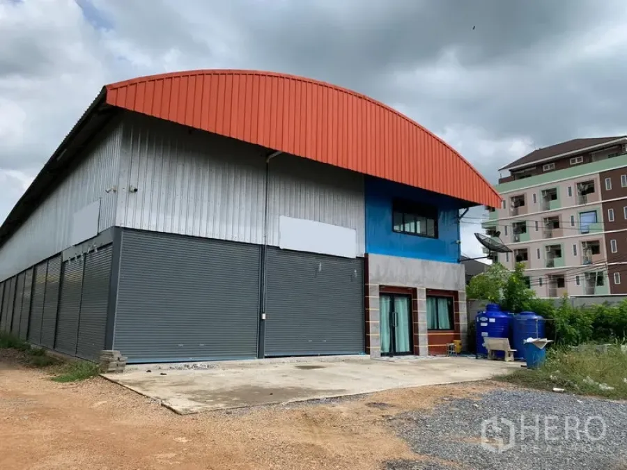 Warehouse for sale in Lam Luk Ka, Pathum Thani - Front exterior of a Lam Luk Ka warehouse with red arched roof, multiple gray roller shutters, attached office, blue water tanks, and a concrete loading yard.