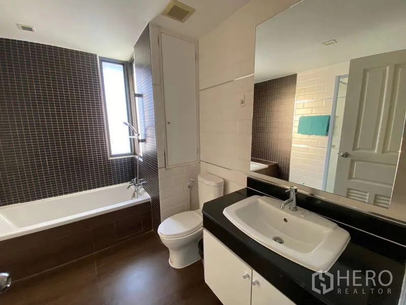 Condominium for rent in Khlong Toei, Bangkok - Well-lit bathroom with window, bathtub with shower, large mirror and white vanity.