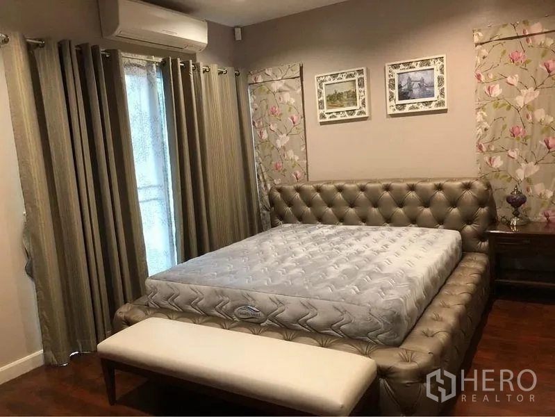 Detached House for rent or sale in Prawet, Bangkok - Air-conditioned bedroom with tufted bed, bench, and floral wall accents.