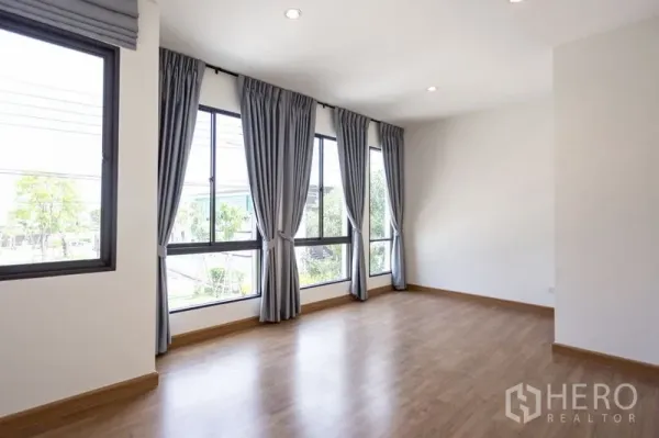 Townhouse for rent in Bang Phli, Samut Prakan - Spacious room with corner floor-to-ceiling windows, grey curtains and wood laminate flooring.
