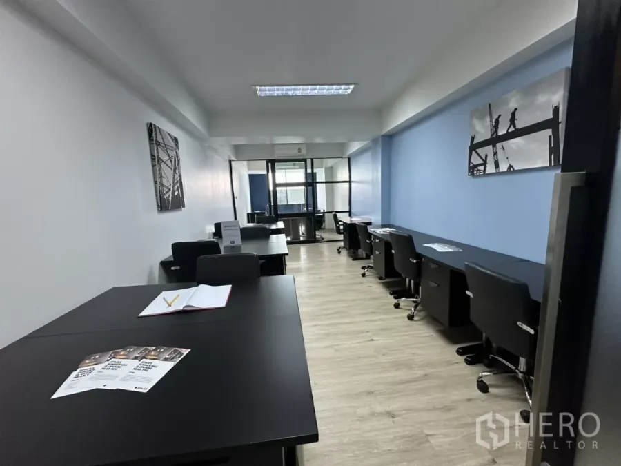 办公空间 出租在Watthana, Bangkok - 开放式办公区，成排黑色书桌与办公椅，蓝色点缀墙与木纹地板，尽头为玻璃会议室。