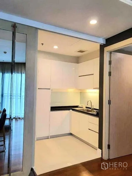 Condominium for rent or sale in Khlong Toei, Bangkok - Open-plan view to a white fitted kitchen off the hallway with recessed ceiling lights and wood floors.