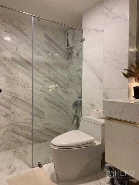 Condominium for sale in Khlong Toei, Bangkok - Second bathroom with white marble walls, glass shower enclosure, vanity and toilet.
