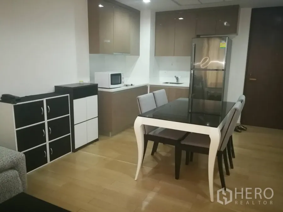 Condominium for rent in Khlong Toei, Bangkok - Open-plan kitchen with glossy cabinets, stainless refrigerator, microwave and a dining table on light wood flooring.
