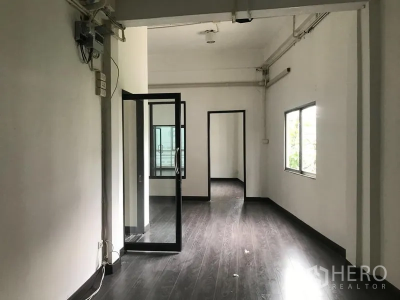 Shophouse for rent in Watthana, Bangkok - Open-plan floor with dark laminate, interior glass door and side window connecting multiple rooms.