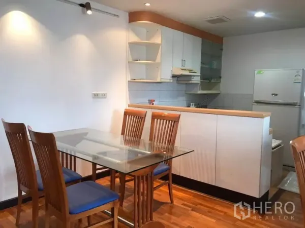 Condominium for rent or sale in Khlong Toei, Bangkok - Glass dining table with wooden chairs beside a compact white kitchen and refrigerator.