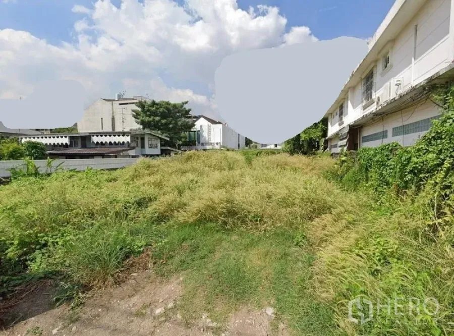 地 出售在Phra Khanong, Bangkok - 曼谷城市低层建筑之间的空地地块，杂草丛生，晴空白云。