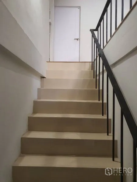 Shophouse for rent in Phra Khanong, Bangkok - Indoor staircase with beige steps and black metal handrail leading to a white door.