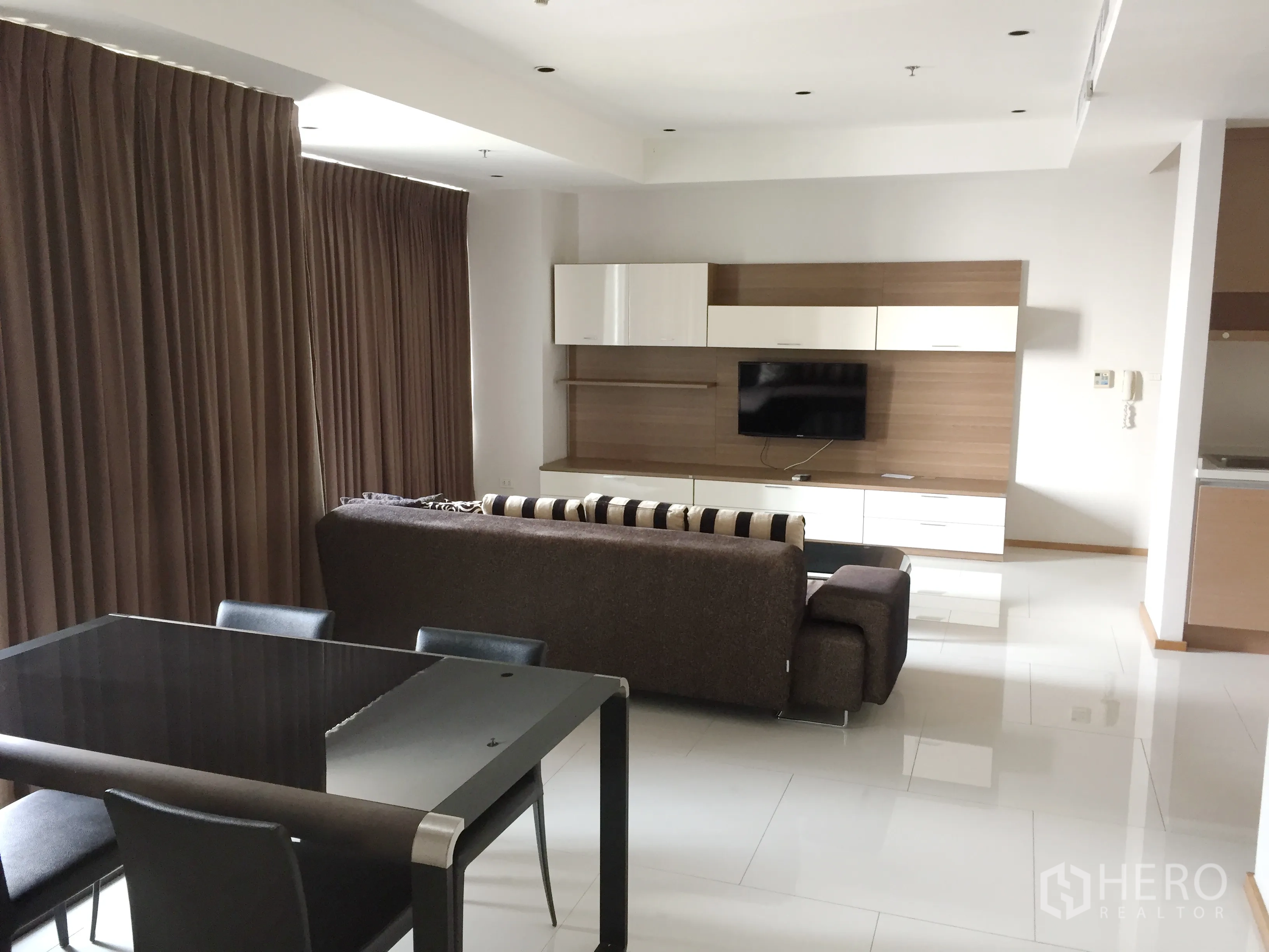 Condominium for rent in Khlong Toei, Bangkok - Open-plan living and dining area with sectional sofa, TV wall unit, and full-length curtains in a modern condo.