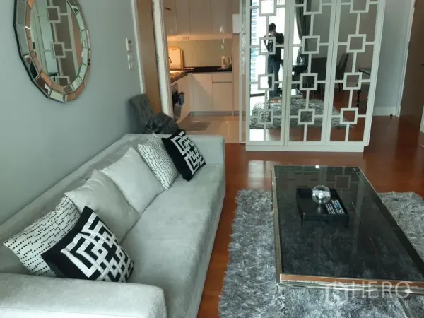 Condominium for rent or sale in Khlong Toei, Bangkok - Contemporary living area facing the kitchen, with mirrored partition, plush rug and wood flooring.
