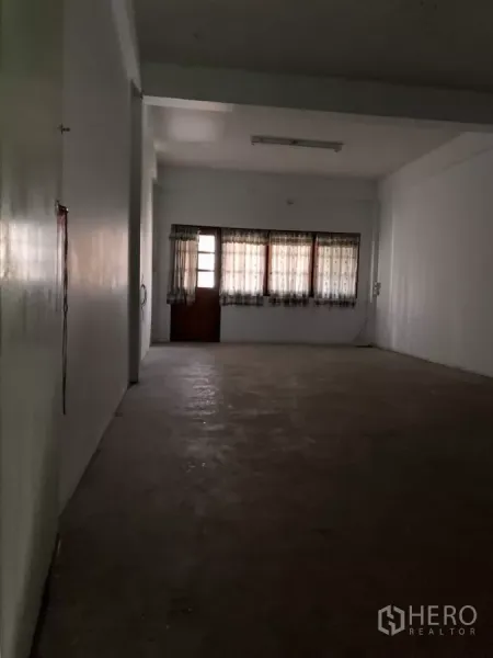 Shophouse for rent in Suan Luang, Bangkok - Deep open-plan floor with concrete surface, curtained windows and a rear wooden door.