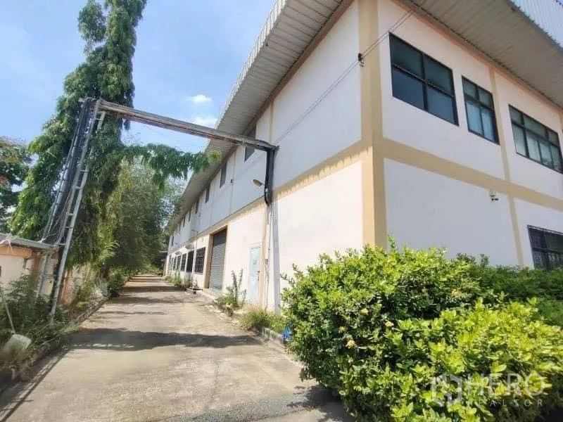 货栈 出售在Khlong Toei, Bangkok - 两层仓库侧立面与金属屋顶，沿侧边车道与绿化，晴天景观。