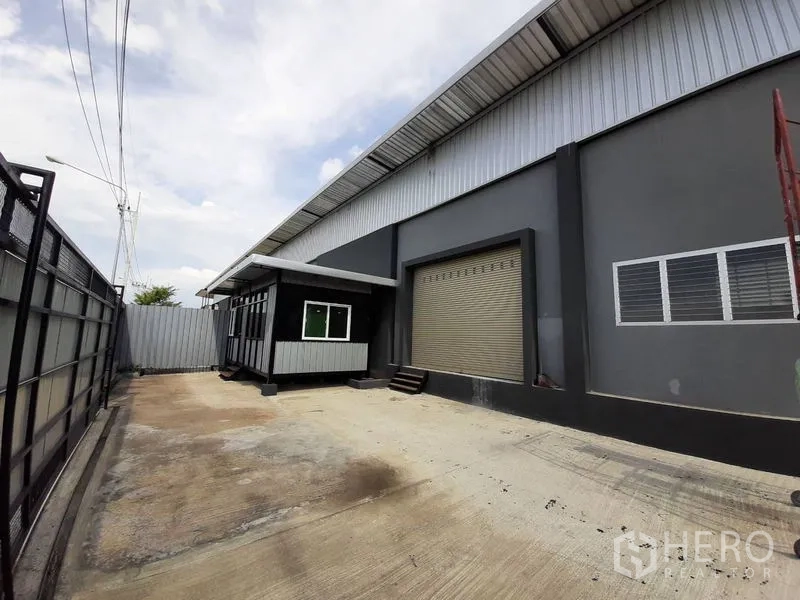 Warehouse for rent in Lam Luk Ka, Pathum Thani - Loading zone with roller shutter, small office booth, and wide concrete yard for trucks.