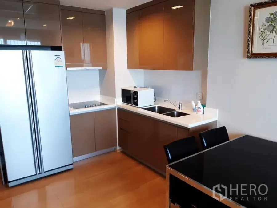 Condominium for rent in Khlong Toei, Bangkok - Modern condo kitchen with glossy cabinets, double-door fridge, cooktop, microwave and sink beside a dining table.